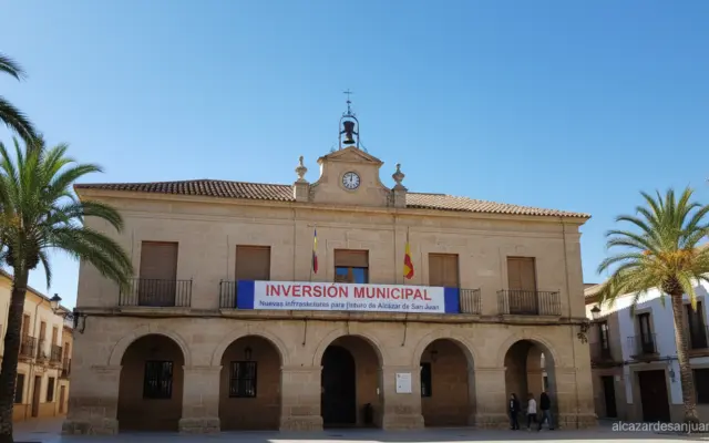 Wide-angle shot of a small-town Spanish town hall (ayuntamiento) facade with a clear blue sky and a banner announcing muni...